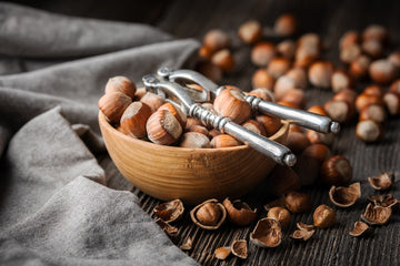 Loads of hazelnuts laying around on top of a wooden table with nutcracker right by their side.